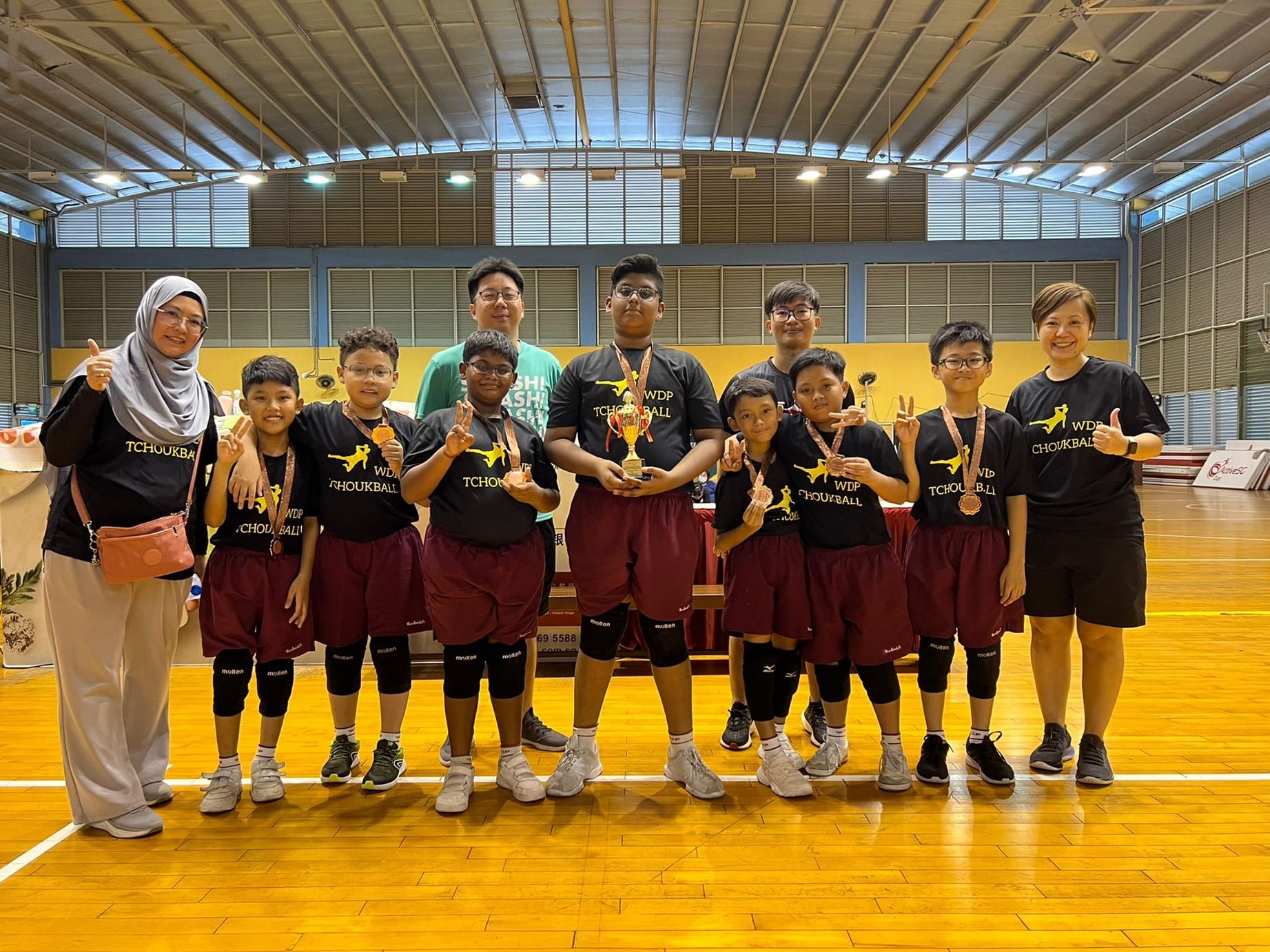 A youth tchoukball team in black WDP shirts poses with a trophy and medals inside an indoor sports hall.